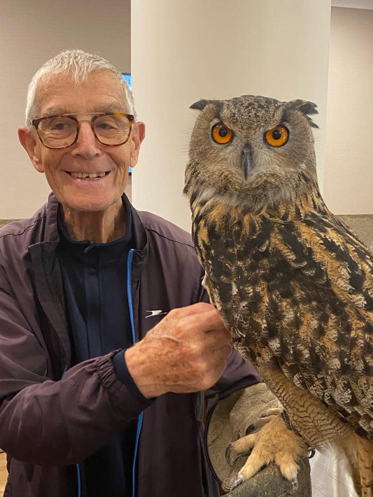A image of a old man in glasses is smiling holding a Eurasian Eagle Owl